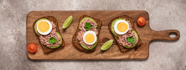 Fototapeta na wymiar Tuna toast. Open sandwich with tuna, rye bread, avocado and boiled egg on a wooden long board, brown grunge background. Top view, flat lay. Banner