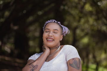 Amidst the greenery of a forest, a nonbinary model of mixed African American and Latinx ancestry poses with a radiant, full smile. Their purple braids add a pop of color.