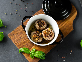 Garlic bulbs Baked with olive oil and spices in black dish. Garlic roasted, black pepper and basil on wooden board. Black background