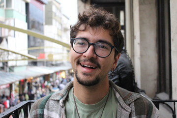 closeup portrait of young man with beard and glasses smiling with crowded city street view *3