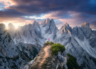 Aerial view of girl on the mountain peak and rocks at colorful sunset in summer. Tre Cime, Dolomites, Italy. Top drone view of woman on trail, cliffs, grass, cloudy sky, sunlight in spring. Hiking - obrazy, fototapety, plakaty