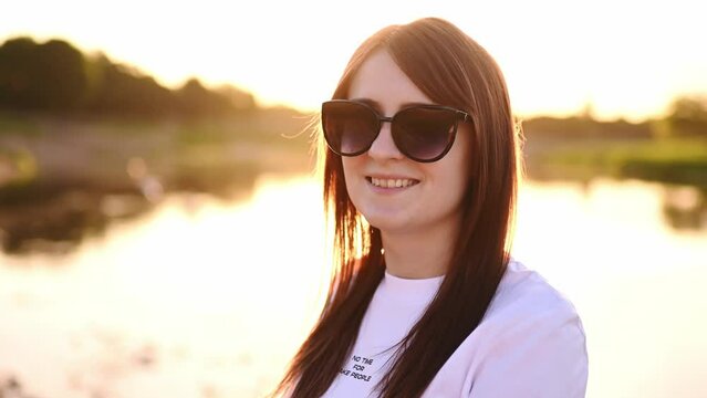 Portrait of a happy woman in sunglasses, white clothes, relaxing at sunset near the water. Summer vacation concept