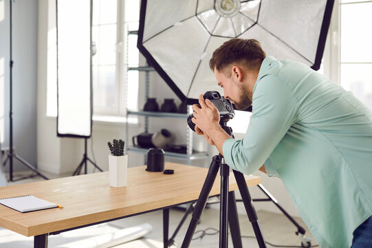 Professional photographer working in his studio workplace. Busy, focused young man standing by tripod with camera and taking photos of little cactus house plant on table. Object photography concept