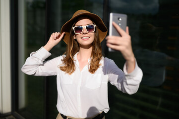 Young happy woman in summer clothes, sunglasses, straw hat posing on a natural background, taking a selfie portrait.