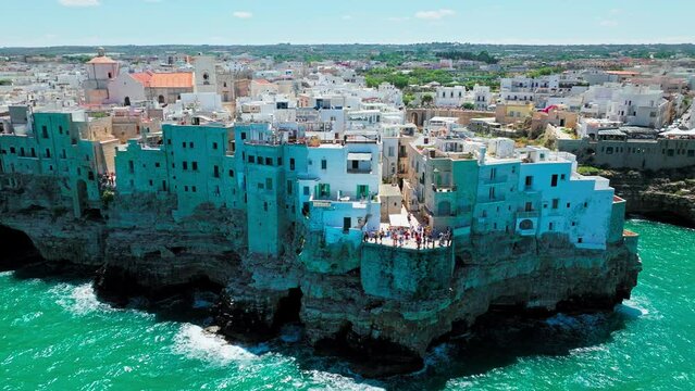 A View From Above Of Tourists Exploring Polignano A Mare Historical Town, Italy. Aerial View Of People Enjoying Balcony Views Facing Adriatic Sea And Walking Along Whitewashed Houses On Rocky Seashore