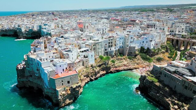 Lama Monachile Cove Beach with clear turquoise water by dramatic rugged cliffs. Aerial drone view of Polignano a Mare with tourists relaxing enjoying and having good time in summer by the beach, Italy