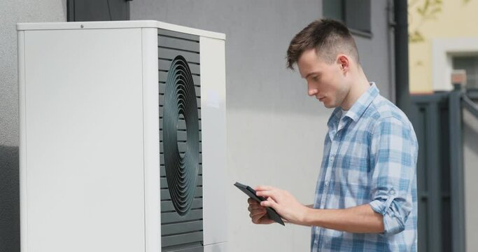A young engineer sets up a heat pump near a private house. Uses a tablet