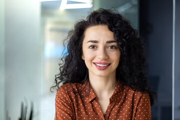Close up photo portrait of beautiful Latin American woman with curly hair , businesswoman inside office building smiling and looking at camera. - obrazy, fototapety, plakaty