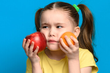 Fototapeta na wymiar On a blue background, the girl holds an apple and a mandarin next to her cheek.