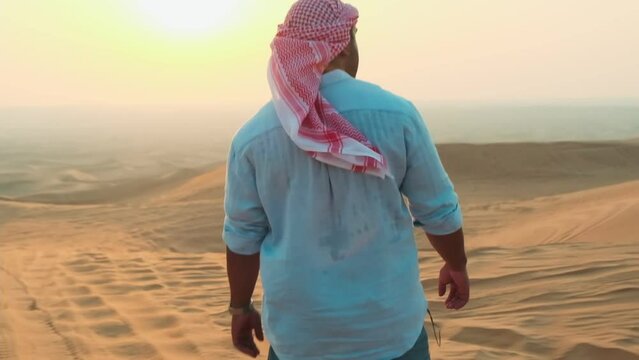A young man in shorts, a shirt and an Arab headscarf walks barefoot on the sand of the desert in the United Arab Emirates