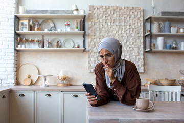 Upset young muslim woman in hijab at home in kitchen looking at phone, checking account, received...