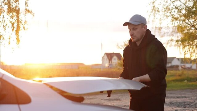 Young man is opening the hood of a car outdoors in the evening on sunset. Driver checking or fixing the automobile engine on the road after a car breakdown. Vehicle service concept. Slow motion.