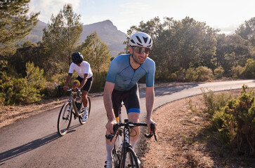 Cyclists in full cycling gear and helmets ride their road bikes on mountain road at sunset. Sportsmen training hard on bicycle outdoors. Spain