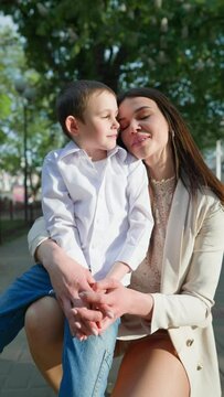 Vertical Video, Loving Boy Child Kisses his Beautiful Mother on Cheek. Concept of Motherhood. Young Attractive Woman Hugging her Son in a City Park on a Sunny Day. Slow Motion.