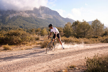 Male cyclist on a gravel bike rides along the gravel trail raising dust from the rear wheel during sunset.Cyclist practicing on gravel road.Spain