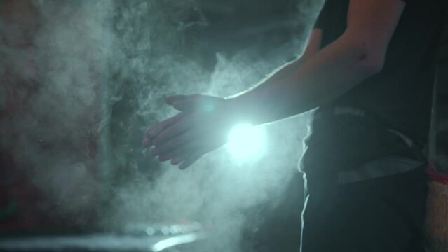 A millennial male rock climber claps his hands with magnesia against a bright lantern during a night workout. Magnesia dust flies out of the hands after the cotton. High quality 4k footage