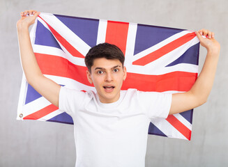 ..Young surprised guy teenager posing with big flag of UK in his hands in studio..