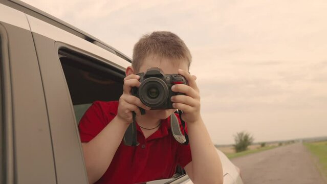 Child boy with camera is sitting in car, taking pictures of nature through window. Childrens journey. Family enjoy car travel on vacation. Photographer child dreams, looks out open car window. Boy kid