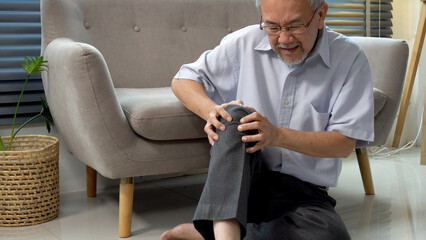 Elderly asian man touching knee hurt painfully problem sitting on floor at home. Patient older...