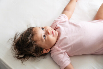 Cheerful adorable daughter wear bodysuit lying down looking curious parent on white bed at home....