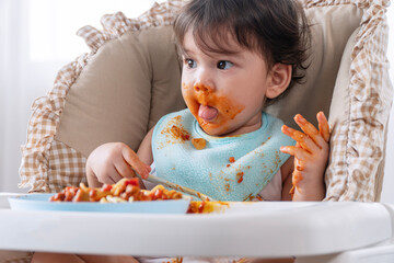 Adorable messy little child girl hungry eating spaghetti spoon sitting in high-powered chair at...