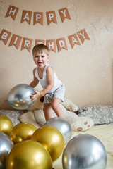 Happy little boy jumping and laughs on bed with balloons in room decorated for birthday celebration. The concept of a birthday