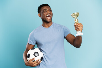 Portrait of overjoyed African American man, soccer player holding ball and trophy cup, celebration success isolated on blue background. Sport victory, winning competition concept