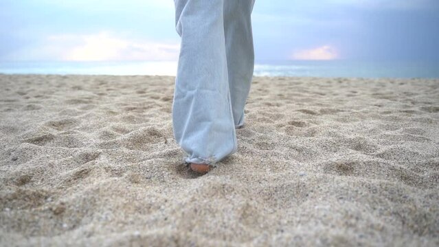 Slender woman's legs walk along the sandy beach toward the sea Wide blue jeans