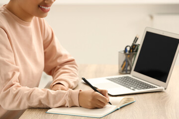 African American woman writing in notepad near laptop at wooden table indoors, closeup