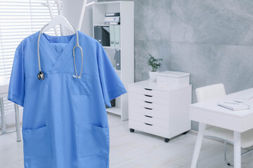 Fototapeta na wymiar Blue medical uniform and stethoscope hanging on rack in clinic. Space for text