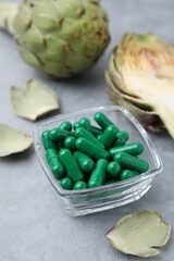 Bowl with pills and fresh artichokes on grey table, closeup