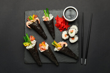 Slate board with tasty sushi cones, rolls, soy sauce, ginger and chopsticks on dark background