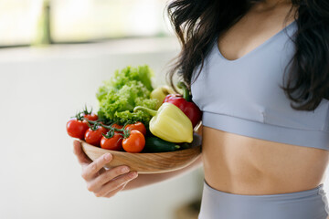 Fit woman holding bowl with fresh organic vegetable standing in modern kitchen, selective focus. Vegan diet, vitamins, healthy lifestyle concept