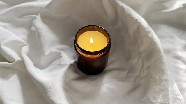 Burning candle in amber glass jar on white cloth, scented candle on linen background