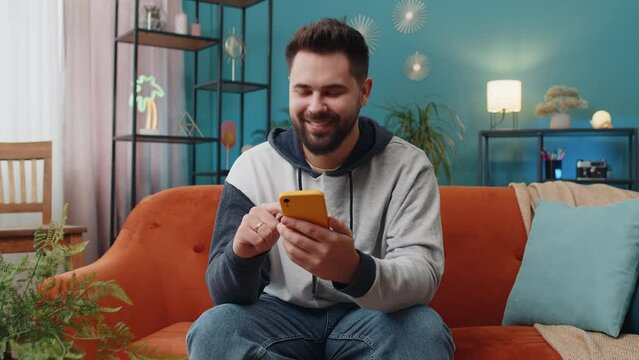 Caucasian man sitting on couch uses mobile phone smiles at home living room apartment. Young guy texting share messages content on smartphone social media applications online, watching relax movie