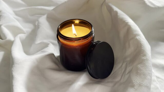 Burning candle in amber glass jar on white cloth, scented candle on linen background