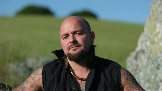 Sitting on a hillside surrounded by rolling grasses, a person in a black vest shows their arm tattoos. A shaved head and goatee express their manliness, and chunky jewelry softens their look.