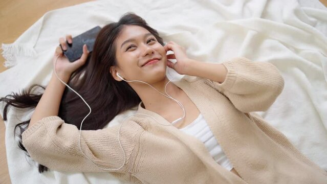 Young asian woman listening to music on the floor in living room at home. Happy asia female using mobile smartphone, wearing headset and lying down on carpet