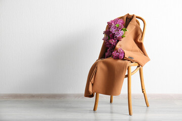 Stylish female coat with beautiful lilac flowers on chair near light wall