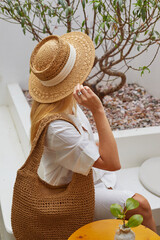 Fashion woman in straw hat with straw bag sitting in modern interior cafe with cup of coffee.