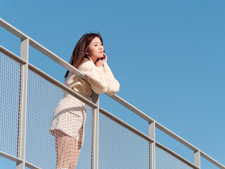 Beautiful young woman with black long hair in white skirt posing on bridge with blue sky background in sunny day. Emotions, people, beauty, travel and lifestyle concept.