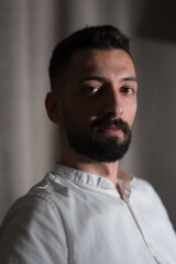 Obraz premium A handsome young Syrian man sits for a portrait in front of a soft, curtained background. He is illuminated by dramatic side lighting.