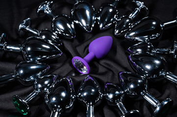 Purple silicone butt plug among many metal ones on a black silk sheet. 