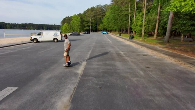 An African American man riding a Onewheel electric skateboard surrounded by lush green trees, plants and grass with parked cars at Proctor Landing Park at lake Acworth in Acworth Georgia USA