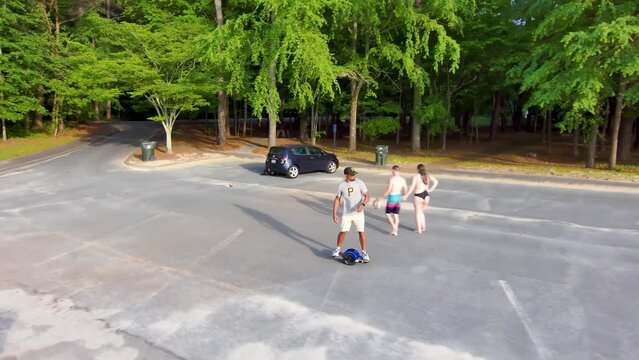 An African American man riding a Onewheel electric skateboard surrounded by lush green trees, plants and grass with parked cars at Proctor Landing Park at lake Acworth in Acworth Georgia USA