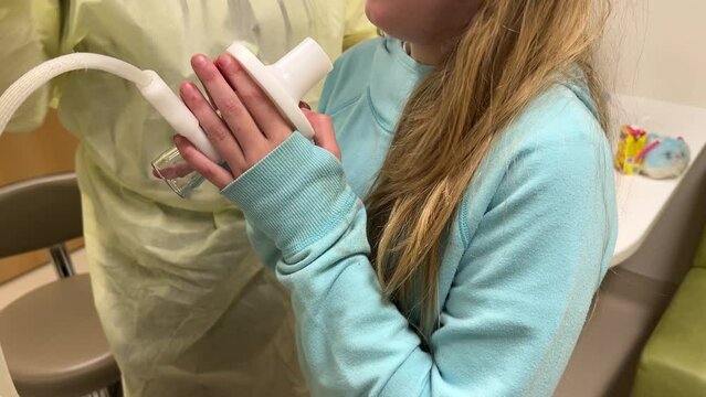 Little girl or long haired boy child kid at doctors office hospital visit to Cystic Fibrosis CF Clinic blowing into respiratory therapy breathing tube with medical technician in ppe.