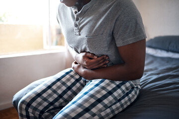 Man, stomach ache and hands holding belly in a bedroom in a home at morning. African male person, sick and indigestion pain in a house on a bed feeling hurt with abdomen cramp and ibs problem - obrazy, fototapety, plakaty