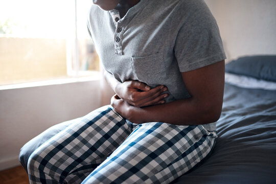 Man, stomach ache and hands holding belly in a bedroom in a home at morning. African male person, sick and indigestion pain in a house on a bed feeling hurt with abdomen cramp and ibs problem