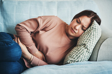 Stomach ache, pain and woman on home sofa with menstrual or period cramps in lounge. Sick, abdomen or colon problem of a female person with hands on tummy for constipation, digestion or virus