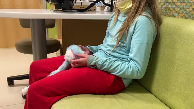 Little girl or long haired boy child kid at doctors office hospital visit to Cystic Fibrosis CF Clinic sitting on green bench couch waiting for doctor and holding plushy toy wearing surgical mask.